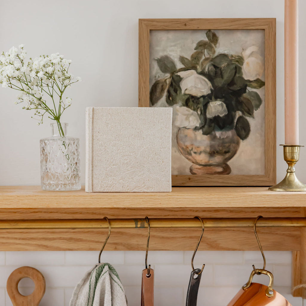 Wooden shelf with copper pots, a cutting board, MUTED + MADE's Little Keepsake Book and a framed floral painting on a white wall.