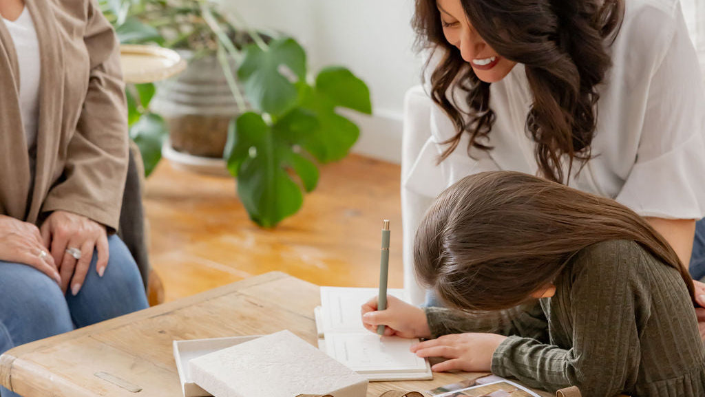 Woman and child sitting at a wooden table with a book, pen, and decorative items in a home setting.