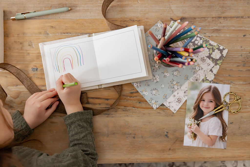 Child coloring a rainbow in a book on a wooden table with art supplies.