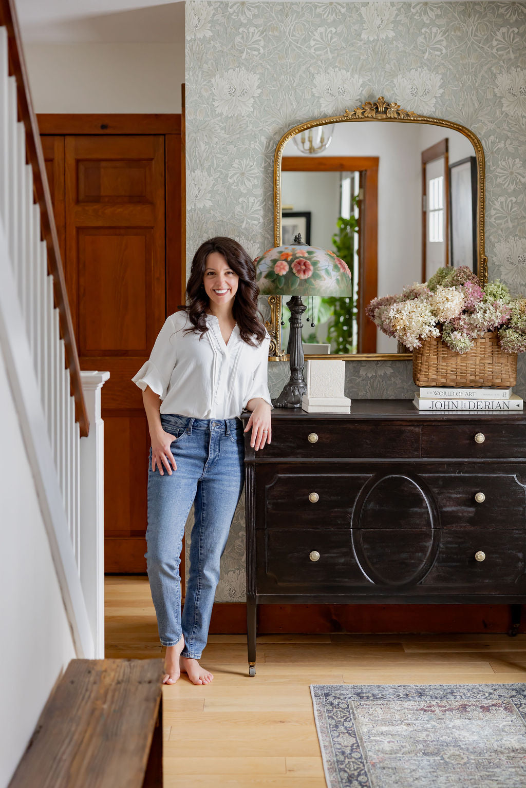 Woman standing in a room with a wooden dresser and decorative mirror.